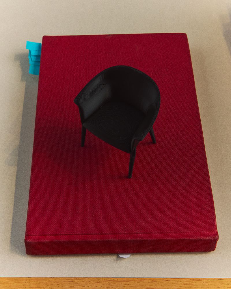 Miniature black chair placed on a red textured book. A small, blue object is clipped to the book's left-side edge, studio setting.