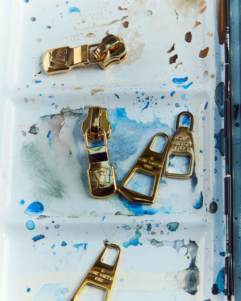 Elevated shot of golden Louis Vuitton zipper pulls arranged on a white and blue watercolor palette, showcasing fashion accessories.