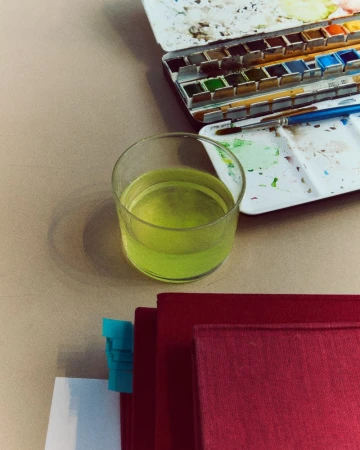 Still life of a glass of water, red books, and a watercolor paint set on a beige surface, showcasing a creative workspace.