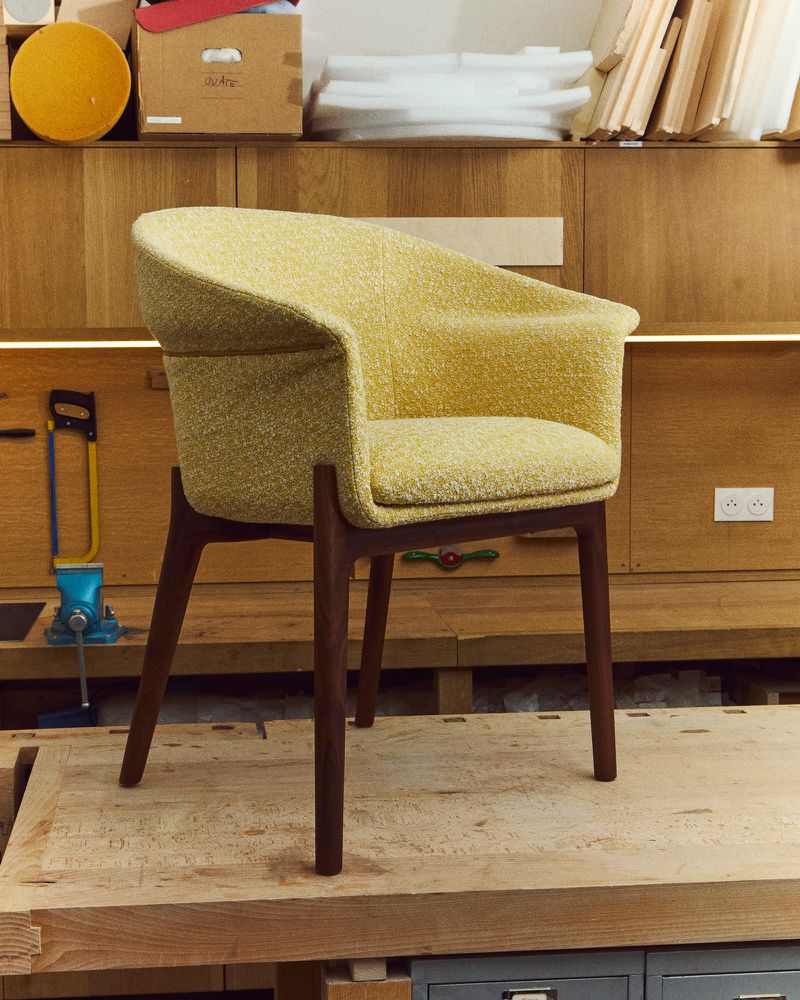 Yellow upholstered armchair with dark wood legs sits on a light wood workbench against wood paneled wall in a workshop.