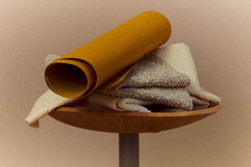 Still life shot featuring a mustard-yellow rolled material stacked on neutral-toned folded textiles, resting on a wooden bowl on a gray stand.