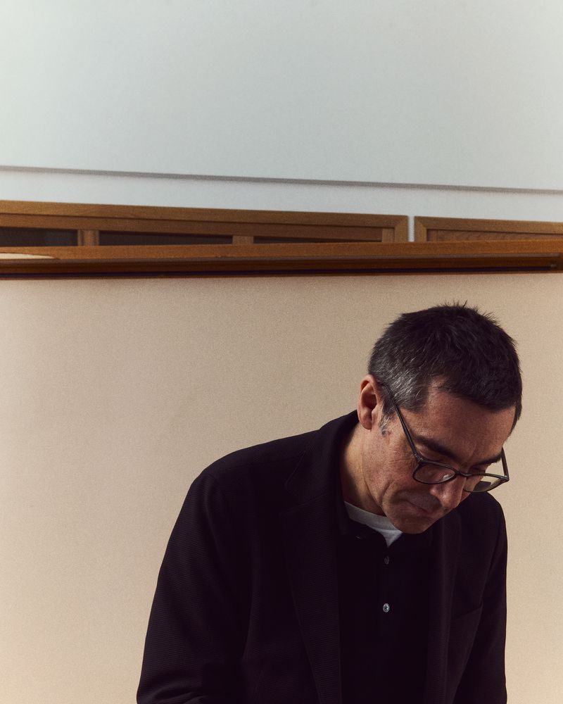 Adult male in glasses, wearing a black jacket, looking down indoors with beige wall and wooden moulding in background, neutral expression.