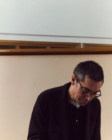 Adult male in glasses, wearing a black jacket, looking down indoors with beige wall and wooden moulding in background, neutral expression.
