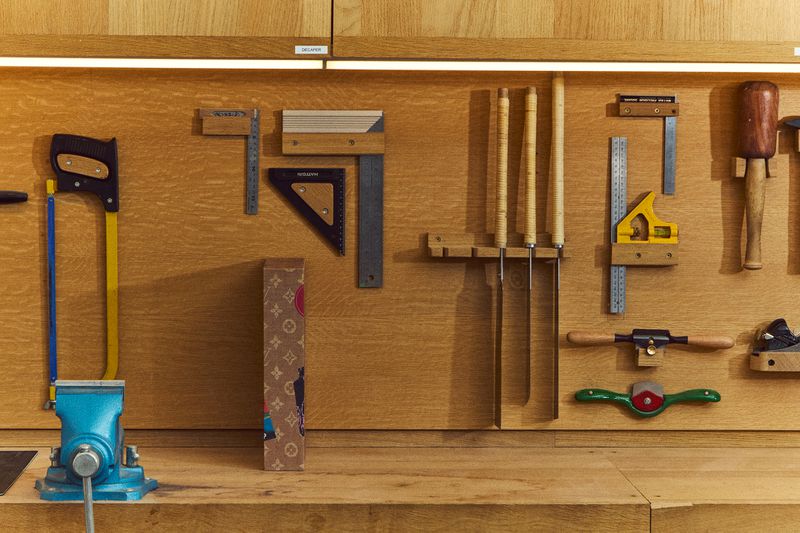 Workbench scene with a blue vise, hand tools and rulers hanging on a wooden wall, and a branded box, with warm lighting.