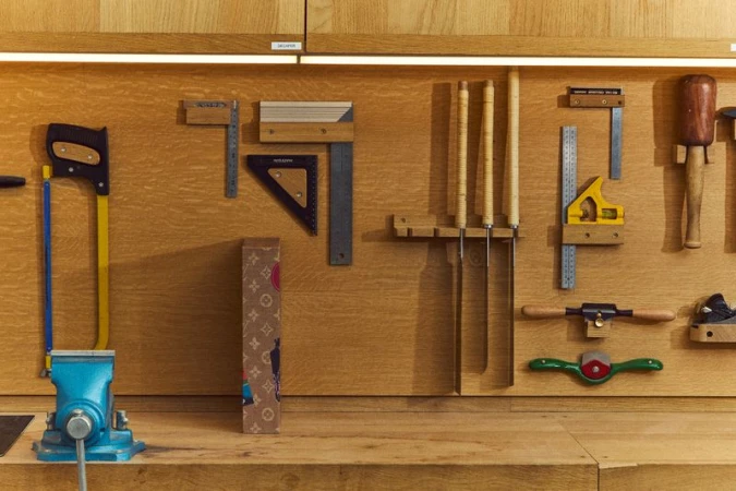 Workbench scene with a blue vise, hand tools and rulers hanging on a wooden wall, and a branded box, with warm lighting.
