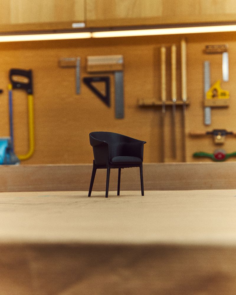 Black chair sits in woodworking shop on wood floor with various tools hanging on the wall in the background under a light.
