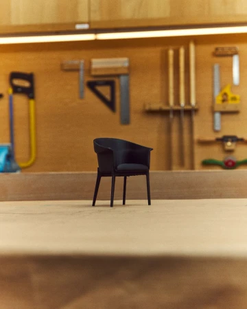 Black chair sits in woodworking shop on wood floor with various tools hanging on the wall in the background under a light.
