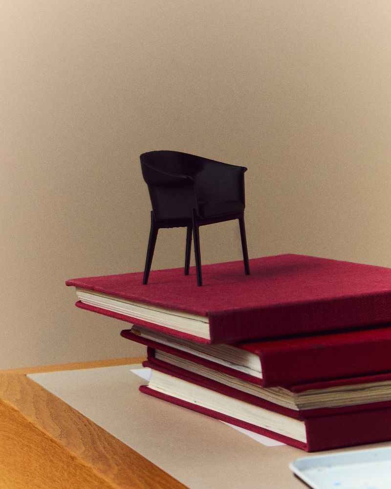 Small black chair sits atop a stack of red books on a wood table, creating a surreal and simple composition against a tan background.