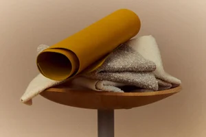 Rolled yellow material and textured bouclé fabric swatches resting on a round wooden stool top against a warm neutral backdrop.