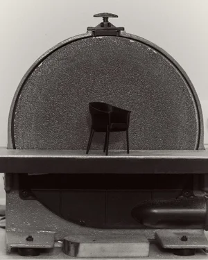 A miniature black designer armchair resting on the metal bed of an industrial disc sander against a pale neutral background.