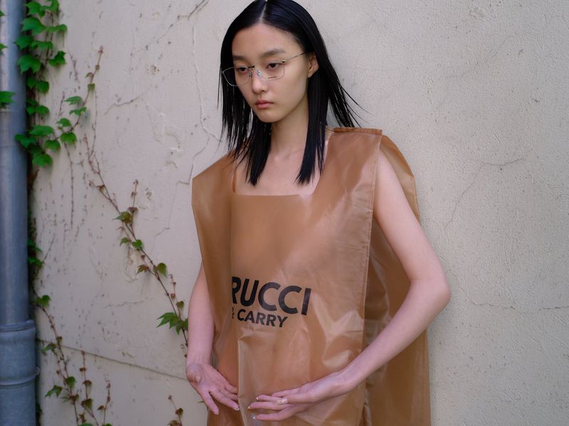 A young Asian woman wears a transparent brown plastic dress with bold black text, standing against a neutral wall with ivy.