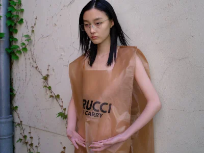 East Asian woman wearing a brown plastic carry bag garment and glasses standing against a textured concrete wall.
