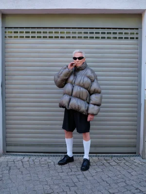 Older man in an oversized metallic puffer jacket and black shorts standing before a closed grey garage door