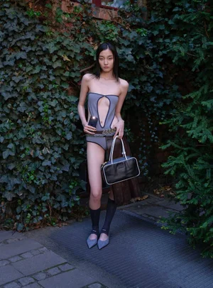 Model standing outdoors against an ivy-covered wall holding a handbag and wearing a cutout bodysuit and heels