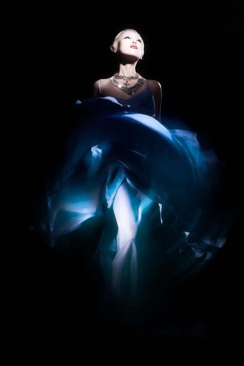 Elegant woman in a blue flowing dress and statement jewelry, illuminated by a spotlight against a dark background, beauty fashion shot.