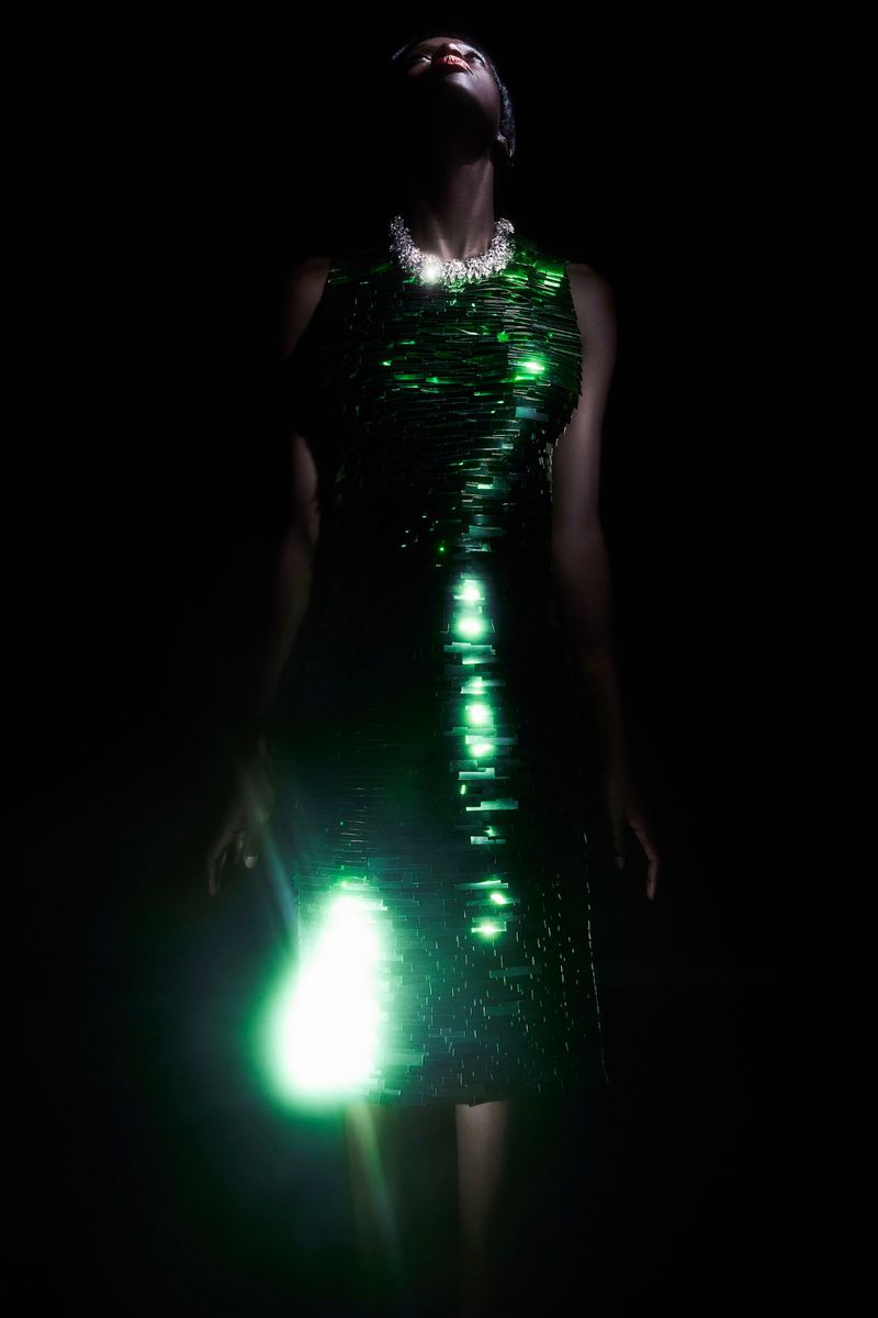 An elegant Black woman in a green sequin dress poses against a dark background, her face illuminated by upward lighting.