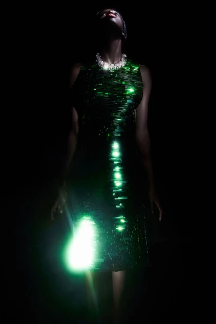 An elegant Black woman in a green sequin dress poses against a dark background, her face illuminated by upward lighting.