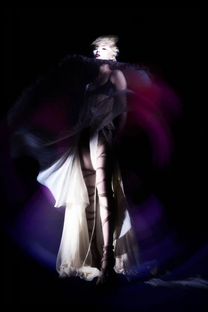 Woman in sheer gown with spiral leg wraps poses in the spotlight with a tulle collar and short blonde hair against a black backdrop.