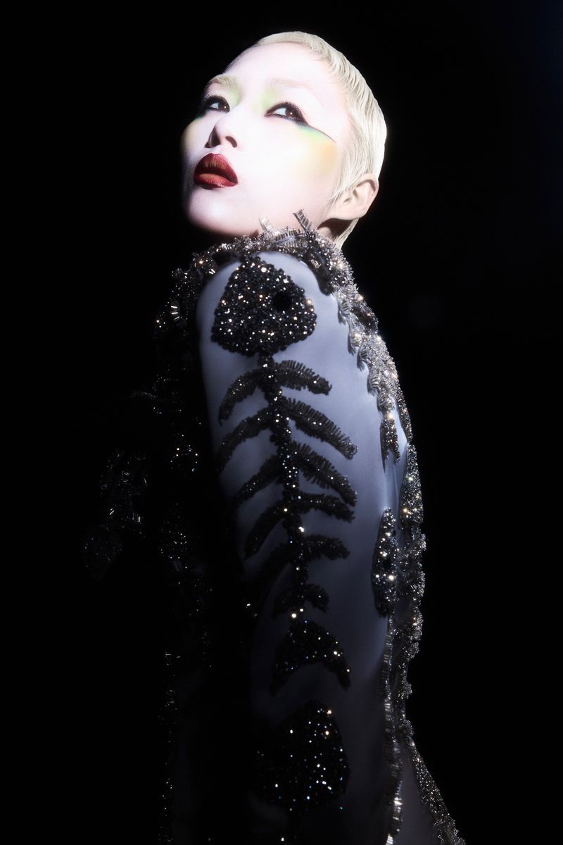 Woman with pale skin and short platinum hair in an embellished grey dress poses in a studio with black background and dramatic lighting.