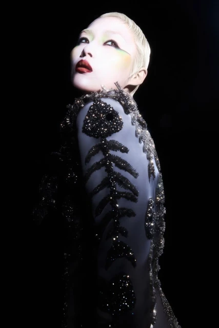 Woman with pale skin and short platinum hair in an embellished grey dress poses in a studio with black background and dramatic lighting.
