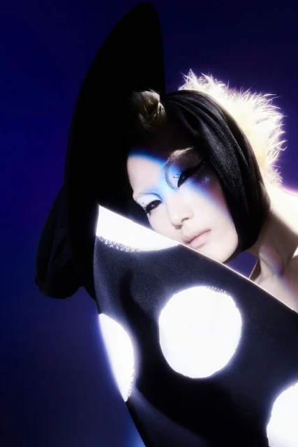 Stylish woman with dramatic makeup poses in front of a blue background, wearing a black hat and polka dot shawl in a studio shot.