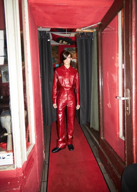 A fashion shot shows a slender person in a red leather jumpsuit standing on a red carpet in an entryway under red ceiling lights.