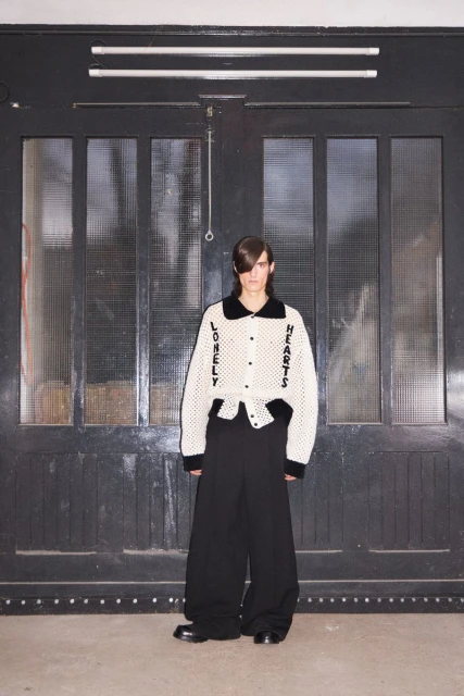 A young adult man stands facing forward in a cream-colored knit cardigan with black trim and wide-legged black trousers against a gray wall.