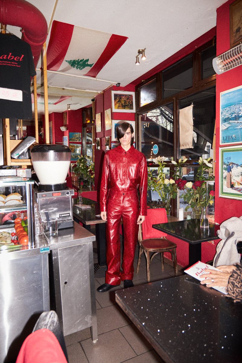 Male model in red jumpsuit stands in a red-toned diner, with a Lebanese flag visible in the background and stainless steel fixtures.