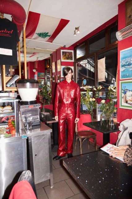 Male model in red jumpsuit stands in a red-toned diner, with a Lebanese flag visible in the background and stainless steel fixtures.