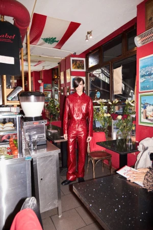 Male model in red jumpsuit stands in a red-toned diner, with a Lebanese flag visible in the background and stainless steel fixtures.