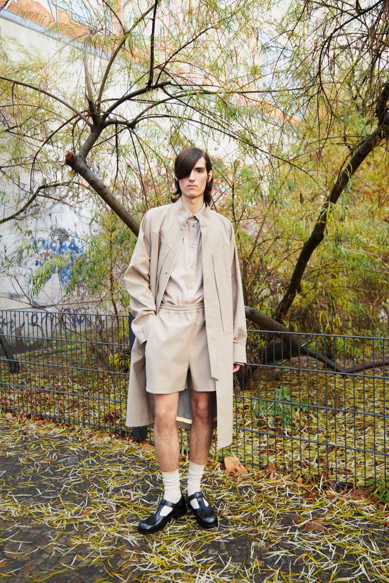 Adult male fashion model poses in a beige shirt, shorts, and trench coat on a path surrounded by foliage in an outdoor setting.