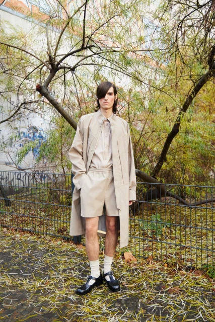Adult male fashion model poses in a beige shirt, shorts, and trench coat on a path surrounded by foliage in an outdoor setting.