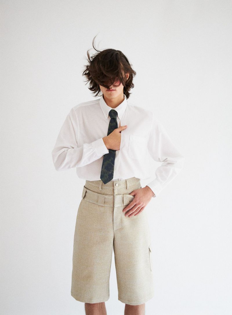 Studio shot of a young adult in a white long-sleeved shirt and beige shorts, styled with a tie, posing against a white background.