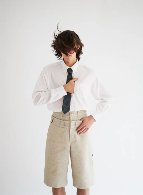 Studio shot of a young adult in a white long-sleeved shirt and beige shorts, styled with a tie, posing against a white background.