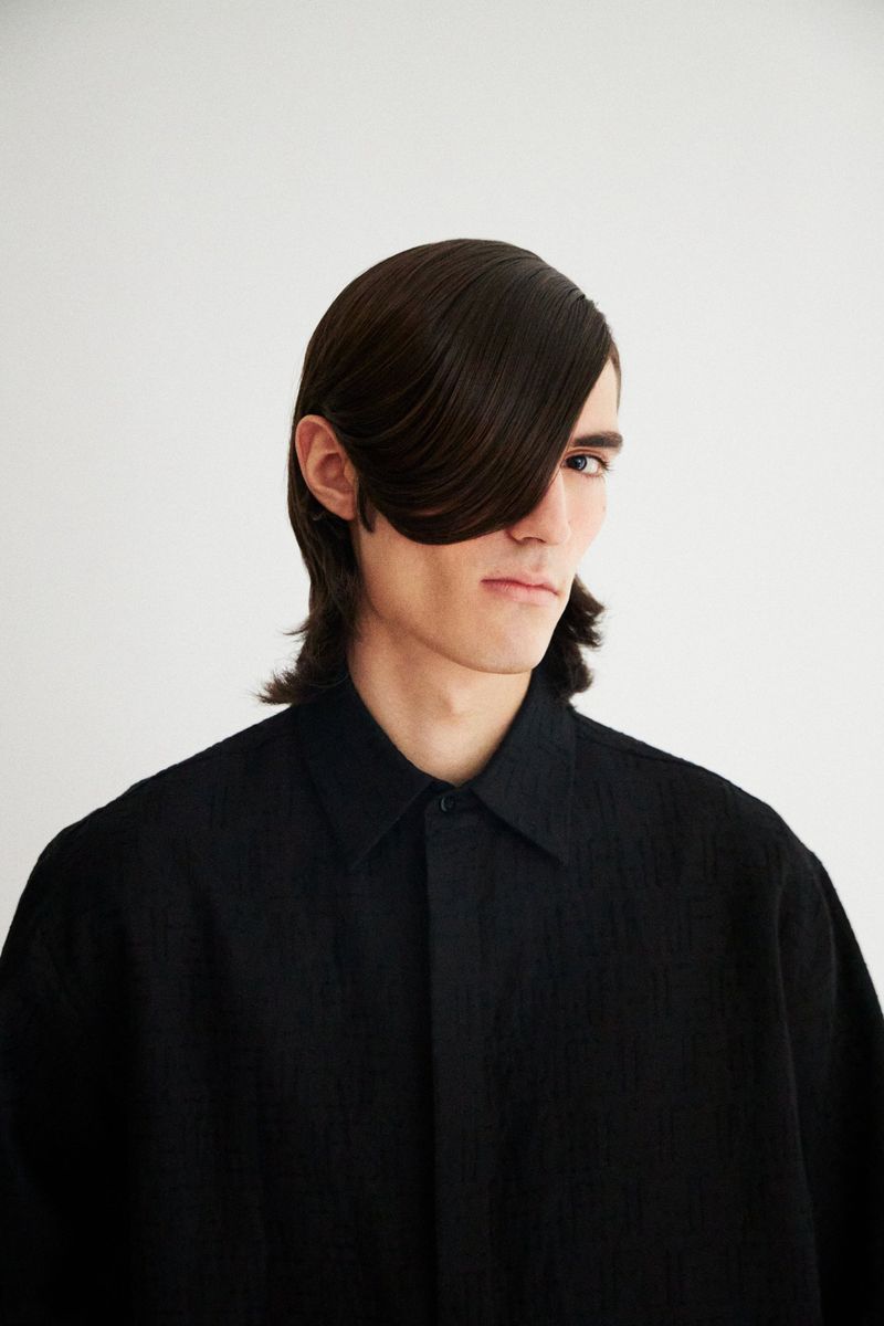 Portrait of a young adult man with long dark hair styled over one eye, wearing a textured black button-down shirt against a white backdrop.