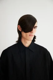 Portrait of a man with dark hair styled across his face wearing a black collared shirt against a white backdrop