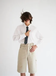 Model with long dark hair covering eyes wearing a white shirt, dark tie, and wide-belted beige shorts against a plain white background.