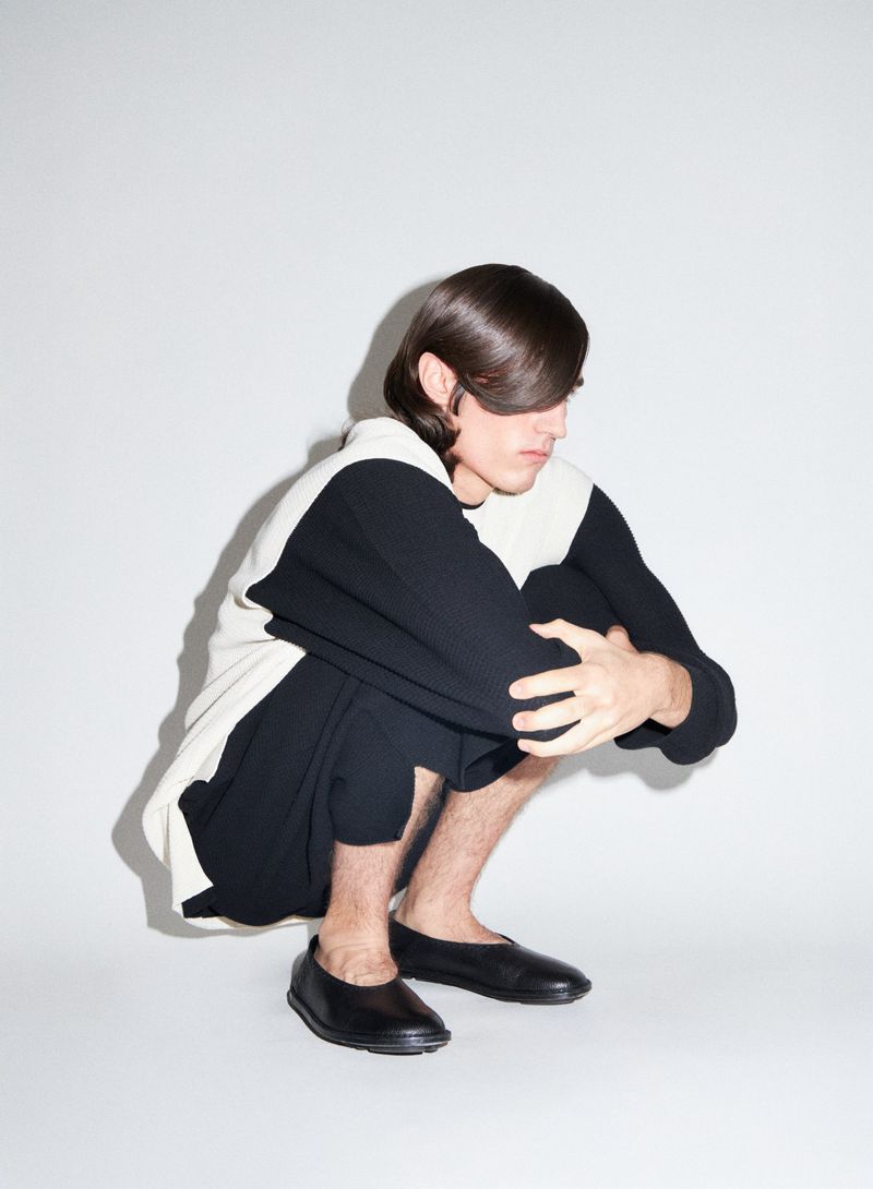 Model crouching with dark hair covering eyes, wearing black and white color-blocked top against a white background