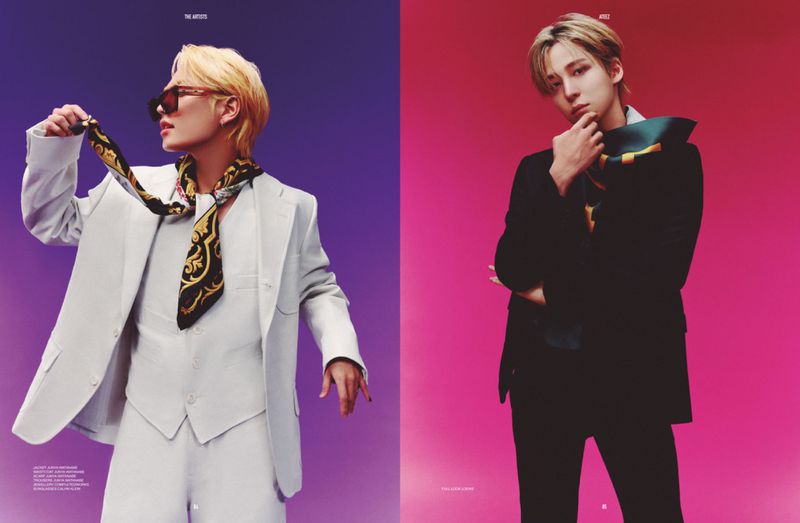 Two stylish men pose in studio: one with blonde hair in a white suit and scarf, the other with brown hair in a black suit and scarf.