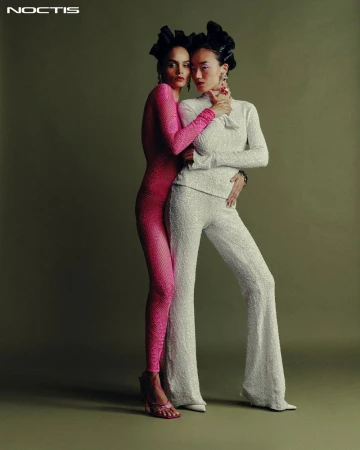 Two multiethnic women in fashionable formalwear, one in pink and one in white, pose in a studio setting. They are wearing elaborate hair accessories.