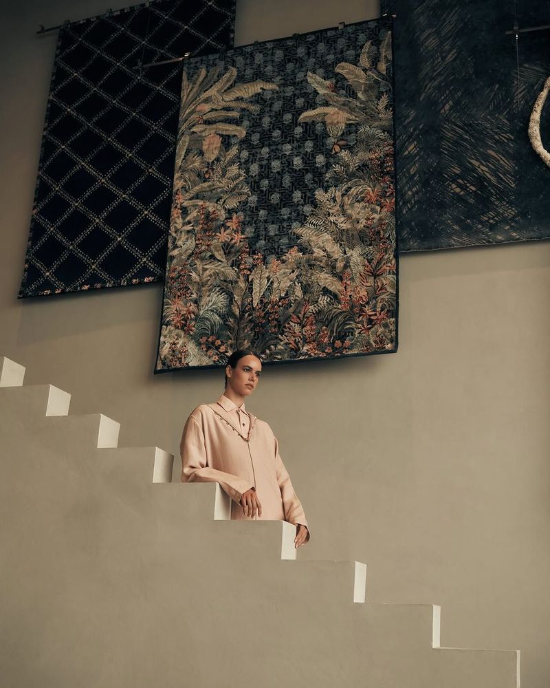 Fashion shot of a young adult female posing on white stairs in front of decorative wall tapestries in an indoor setting, looking off-camera.