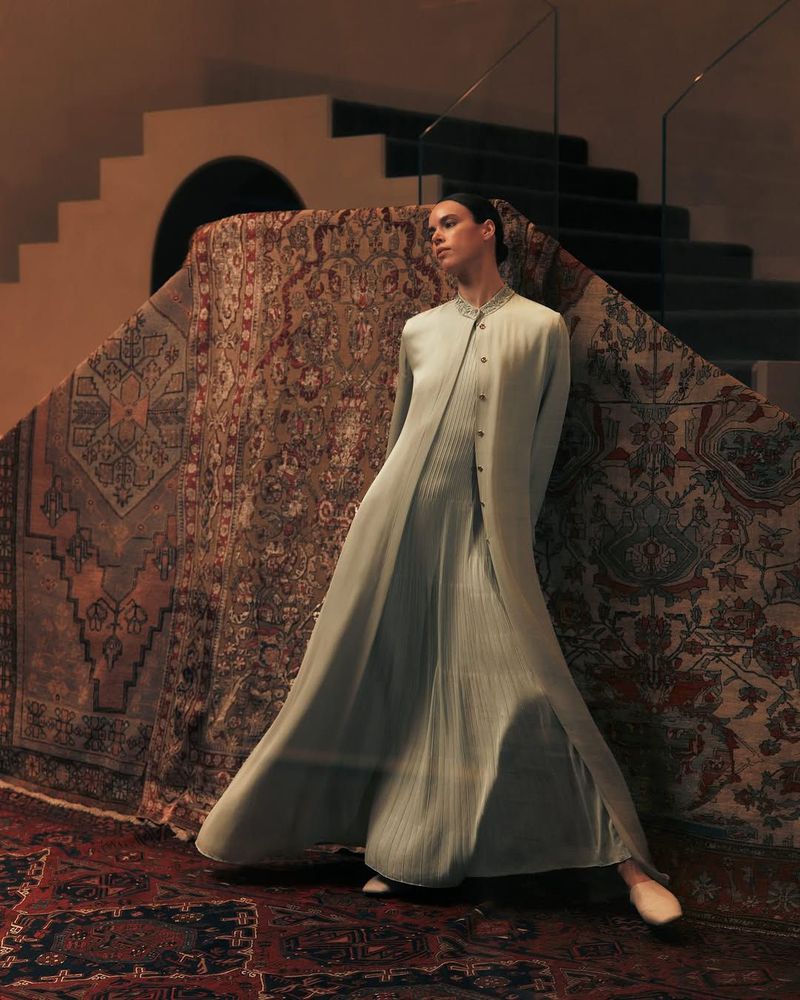 A fair-skinned woman in a pleated dress and coat poses next to a vintage patterned rug in an indoor setting with a staircase background.