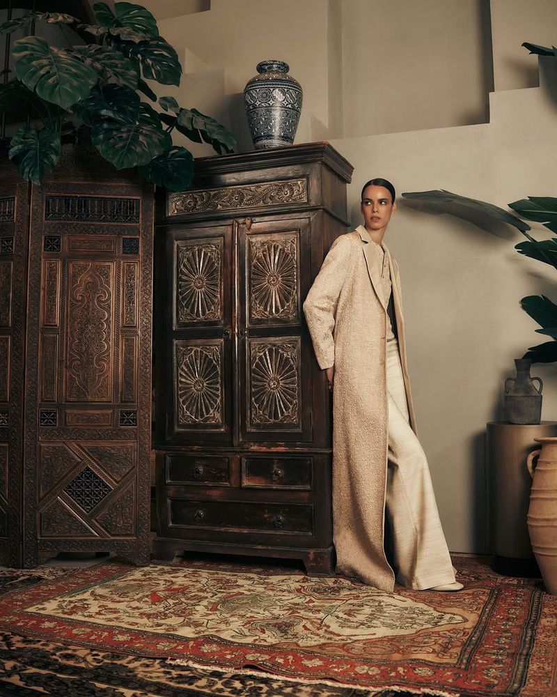 Elegant woman in a long coat and wide-leg pants, standing in an indoor setting with antique furniture and leafy plants, fashion editorial.