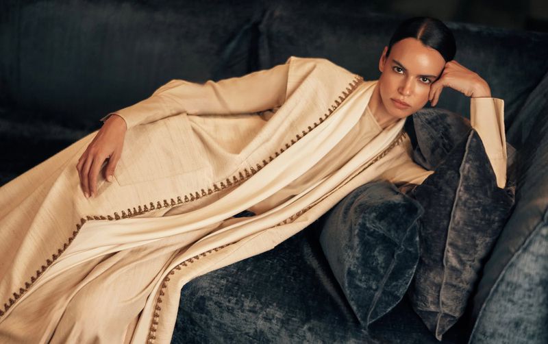 An adult female model is lying on a velvet couch with pillows wearing an off-white long-sleeved dress in an indoor setting.