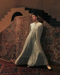 Model wearing a long sage green coat and pleated dress standing against a large draped patterned rug in an interior space