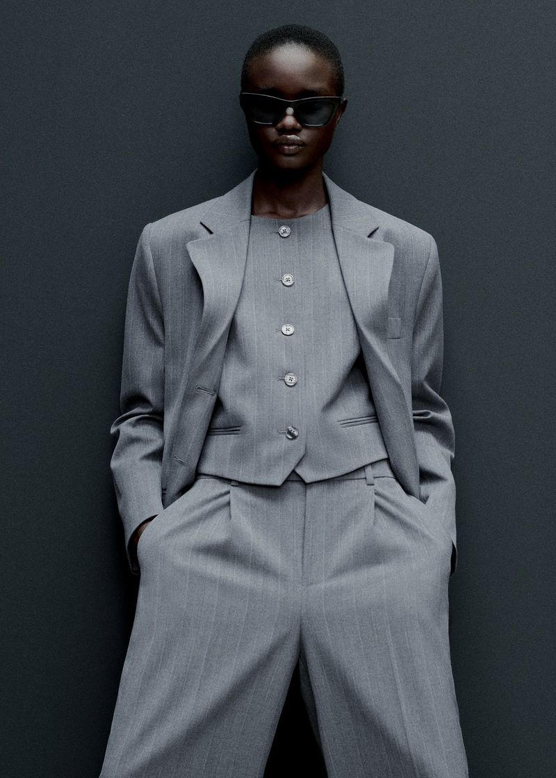 An African-American woman in a pinstripe suit and sunglasses poses against a grey background, showcasing an elegant and modern fashion ensemble.