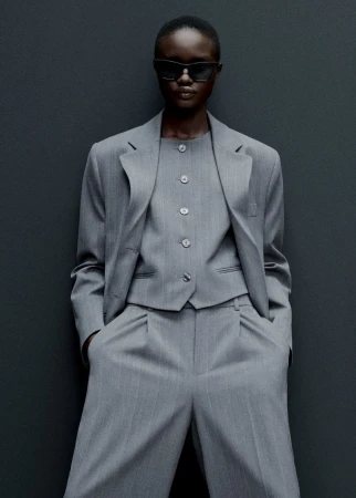 An African-American woman in a pinstripe suit and sunglasses poses against a grey background, showcasing an elegant and modern fashion ensemble.