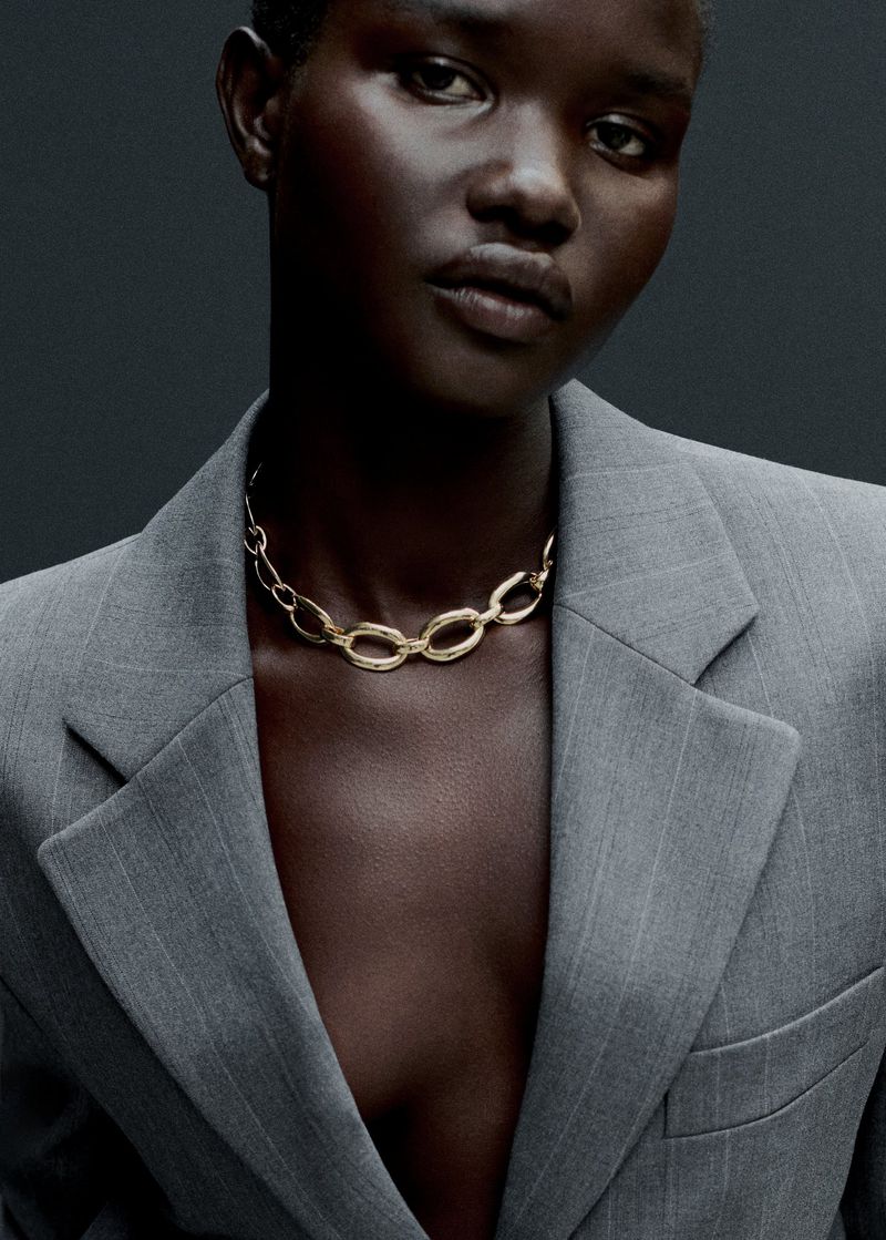 Close-up shot of a dark-skinned model wearing a gray pinstripe blazer and a gold chain necklace against a dark gray background.