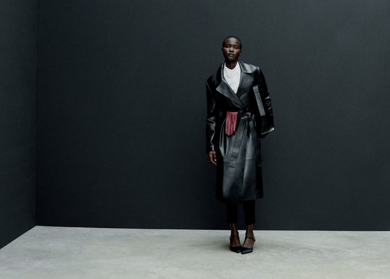 Fashion editorial shot of an adult woman in a black leather trench coat standing against a dark gray wall with a light gray floor.