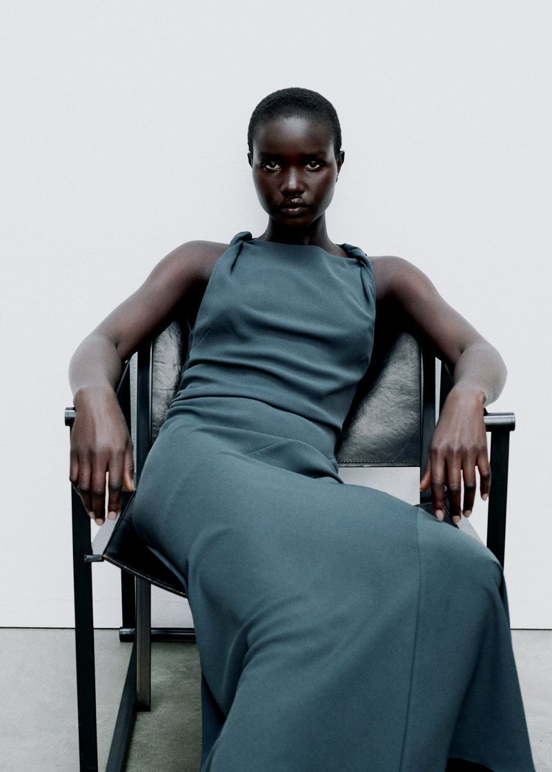 A dark-skinned woman in an elegant teal dress poses in a chair against a neutral background in a fashion setting, capturing a poised look.